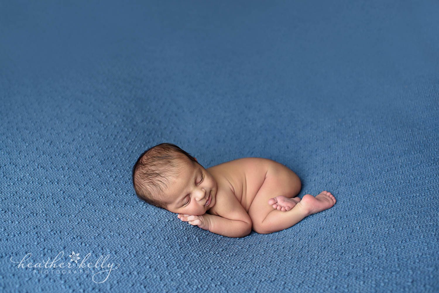 Newborn Photography posing. Sleeping newborn boy on dark blue backdrop with leg tucked under. 