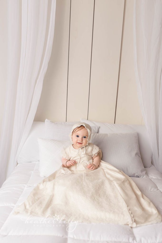 7 month baby girl sitting on bed in baptism gown photo. Baptism portrait photography by ct baby photographer Heather Kelly Photography. 