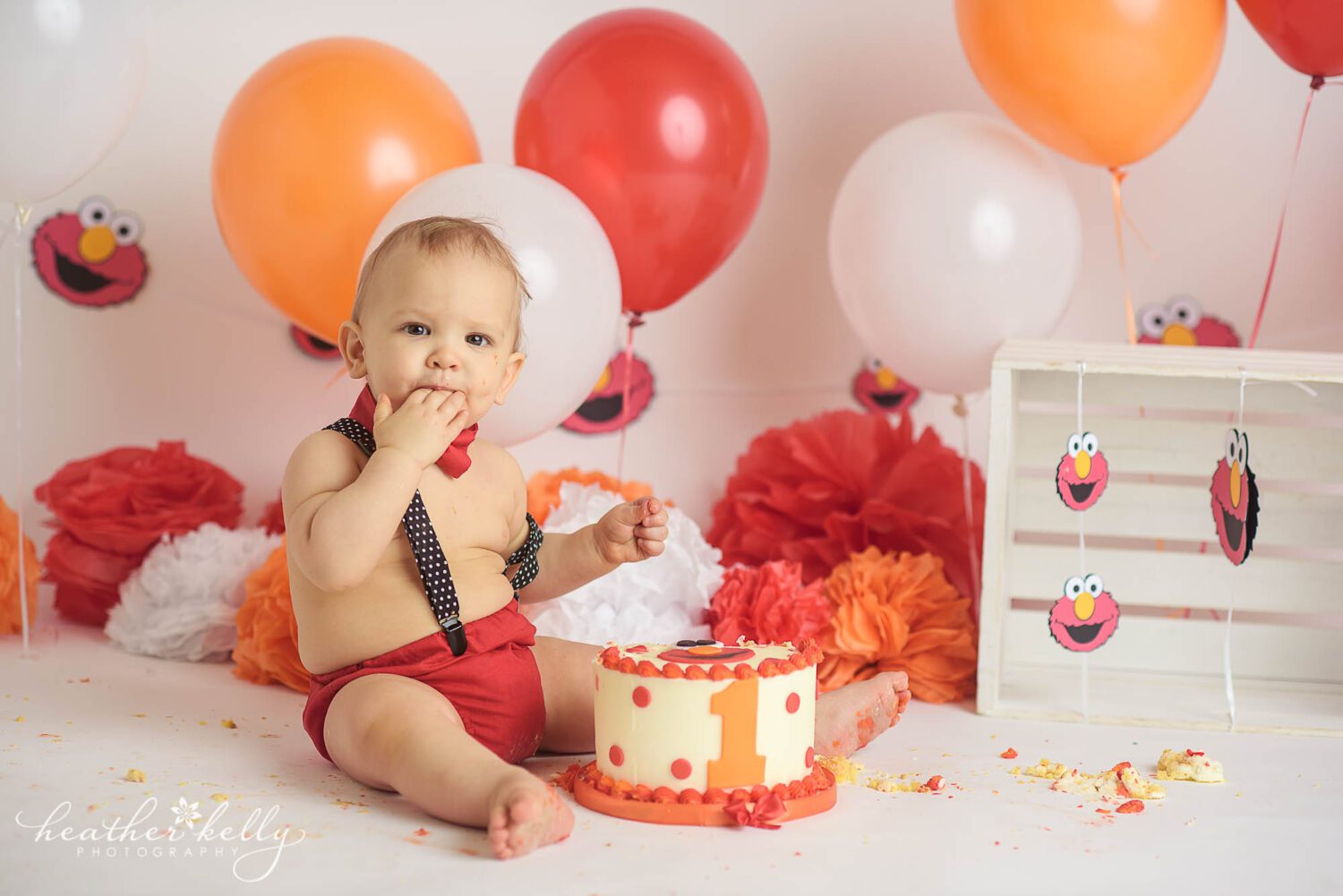 a one year old boy sticks his fingers in his mouth, eating the icing off of his elmo themed cake. The background is decorated in orange, red, and white balloons with an elmo banner. This was his Stratford CT Cake Smash session taken by professional photographer, Heather Kelly Photography