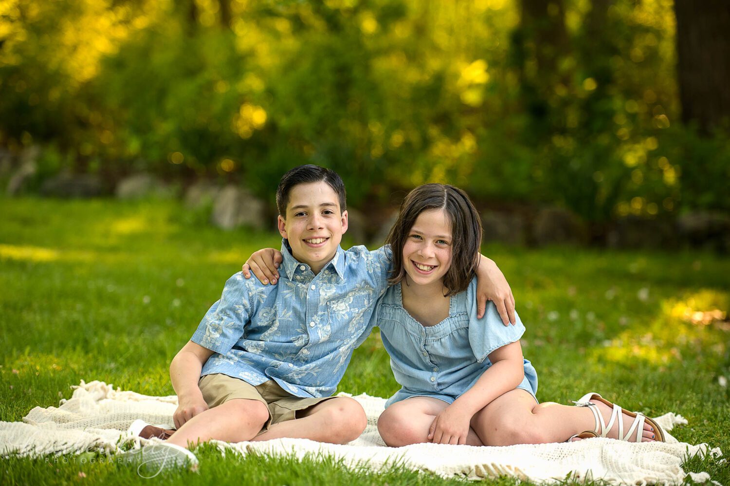 two siblings sitting in the grass on a blanket. They have their arms around each other and are smiling at the camera. 