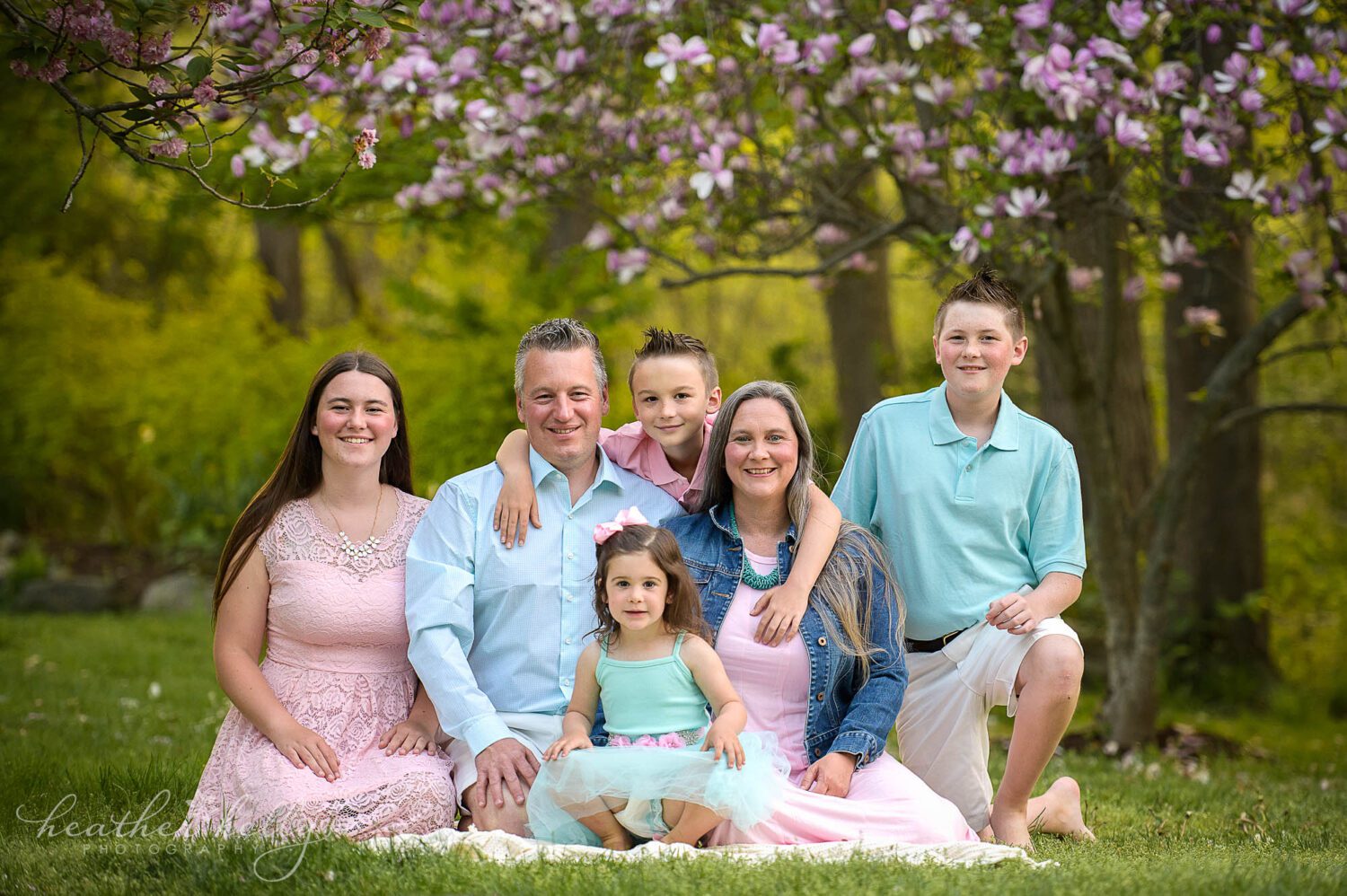 family of 6 in coordinating spring colors sit on a blanket looking at the camera for a springtime portrait session in newtown ct. There is a beautiful pink and purple tree blooming behind them. 