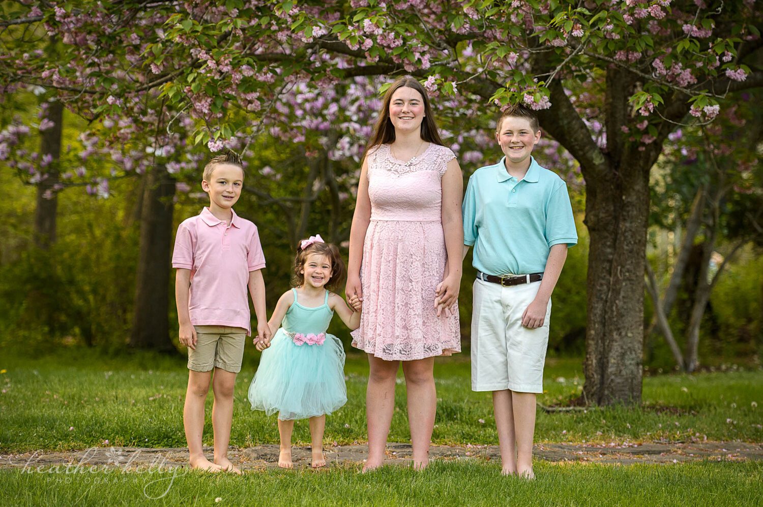 4 kids holding hands and looking at the camera for a spring photography session. There is a beautiful pink and purple tree blooming behind them. 