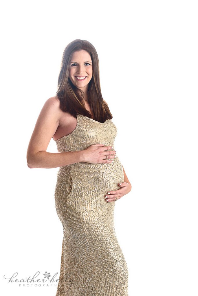 CT studio maternity photo session of a pregnant mom. She is wearing a gold sparkly dress with a completely white background. She is looking and smiling at the camera with her hands above and below her belly. 