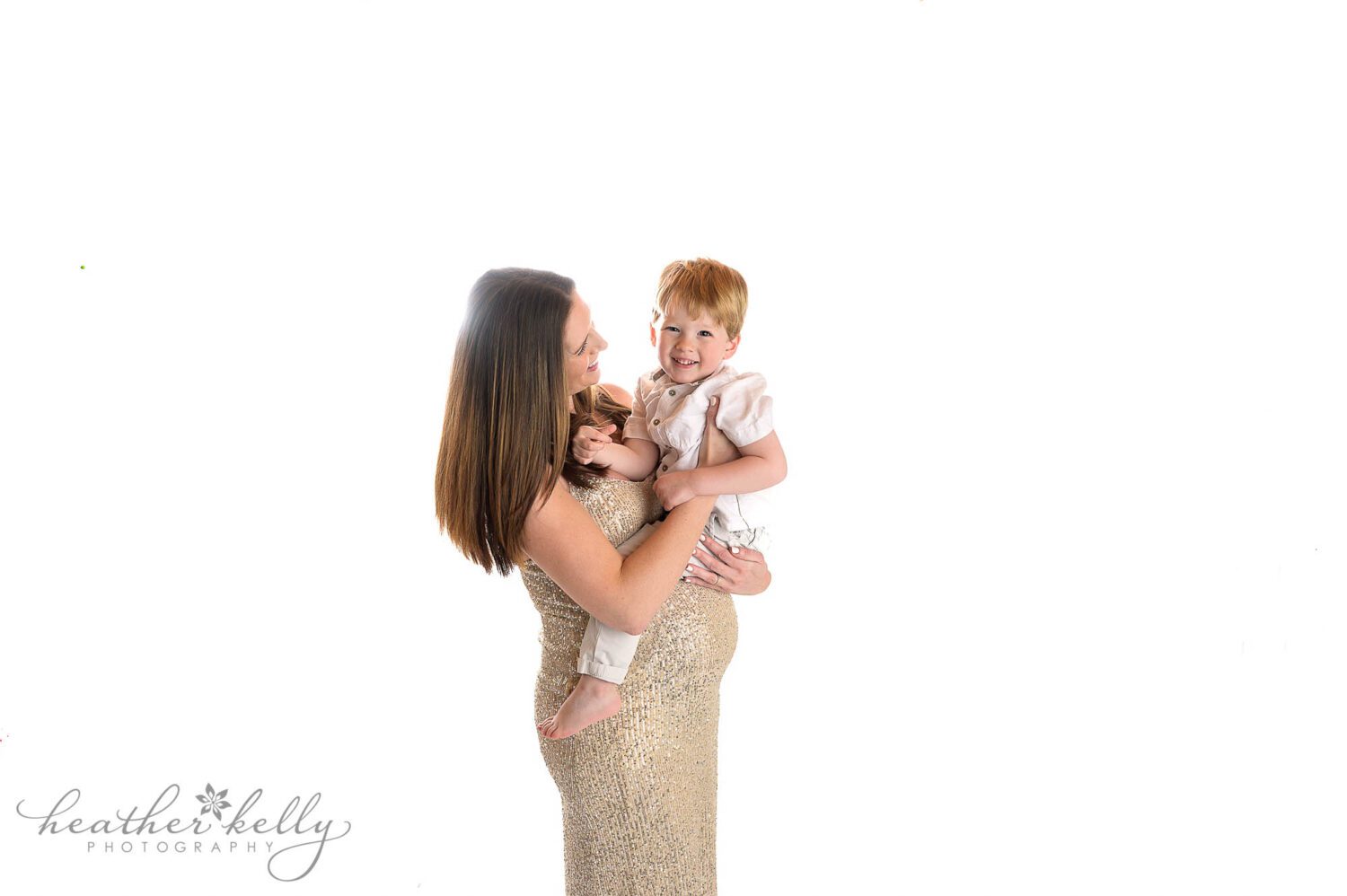 Pregnant mom is posing at her maternity portrait photography session.  There is a bright white background. Mom is in a glittery gold dress and holding her two year old son over her belly. 