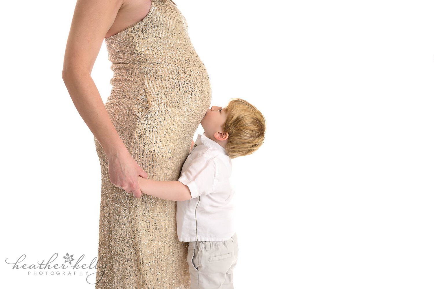 Stamford CT mom to be posing at her maternity portrait photography session.  There is a bright white background. Mom is wearing a sparkly gold dress. Her two year old son is in a white shirt and kissing her belly. 