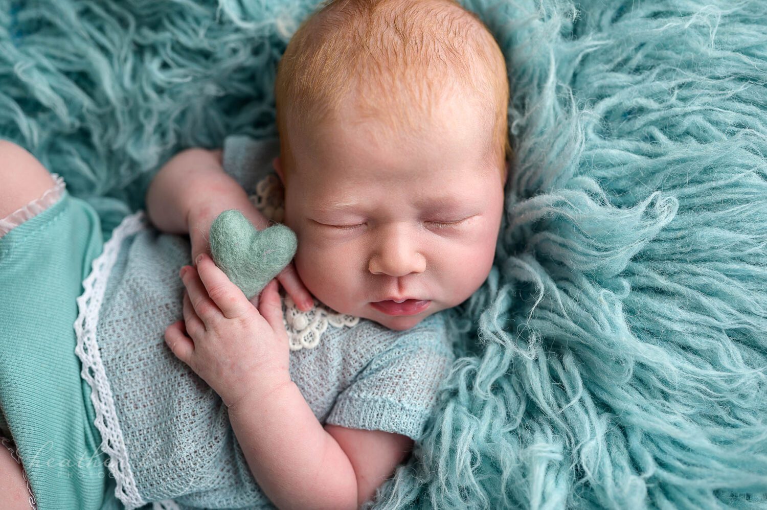 a newborn girl is holding a felted aqua heart. She is posed on a aqua flokati during her newborn session. 