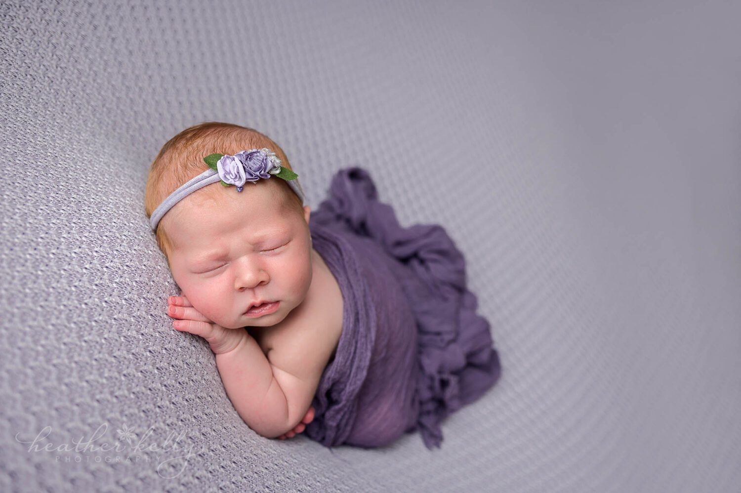 a newborn girl is posed sleeping on a purple backdrop. Her hand is tucked under her chin. A purple wrap is tucked loosely around her. 