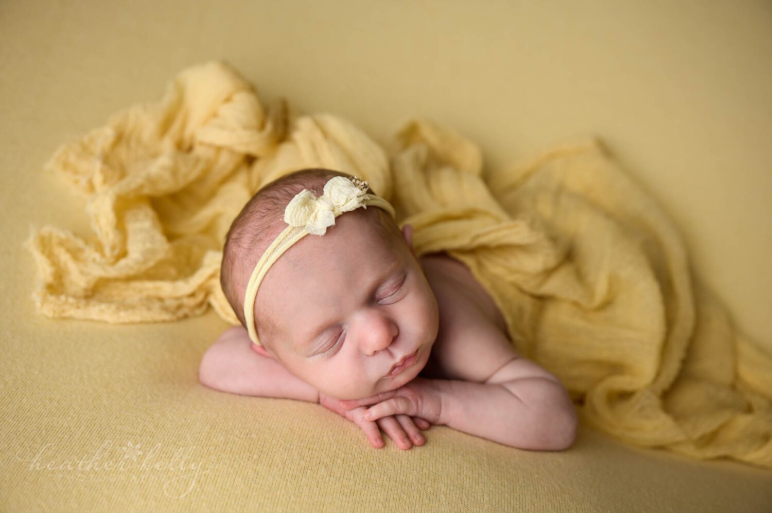 a sleeping newborn girl on a yellow backdrop and a yellow wrap draped loosely on top of her. She is wearing a yellow headband. Newborn photos. 