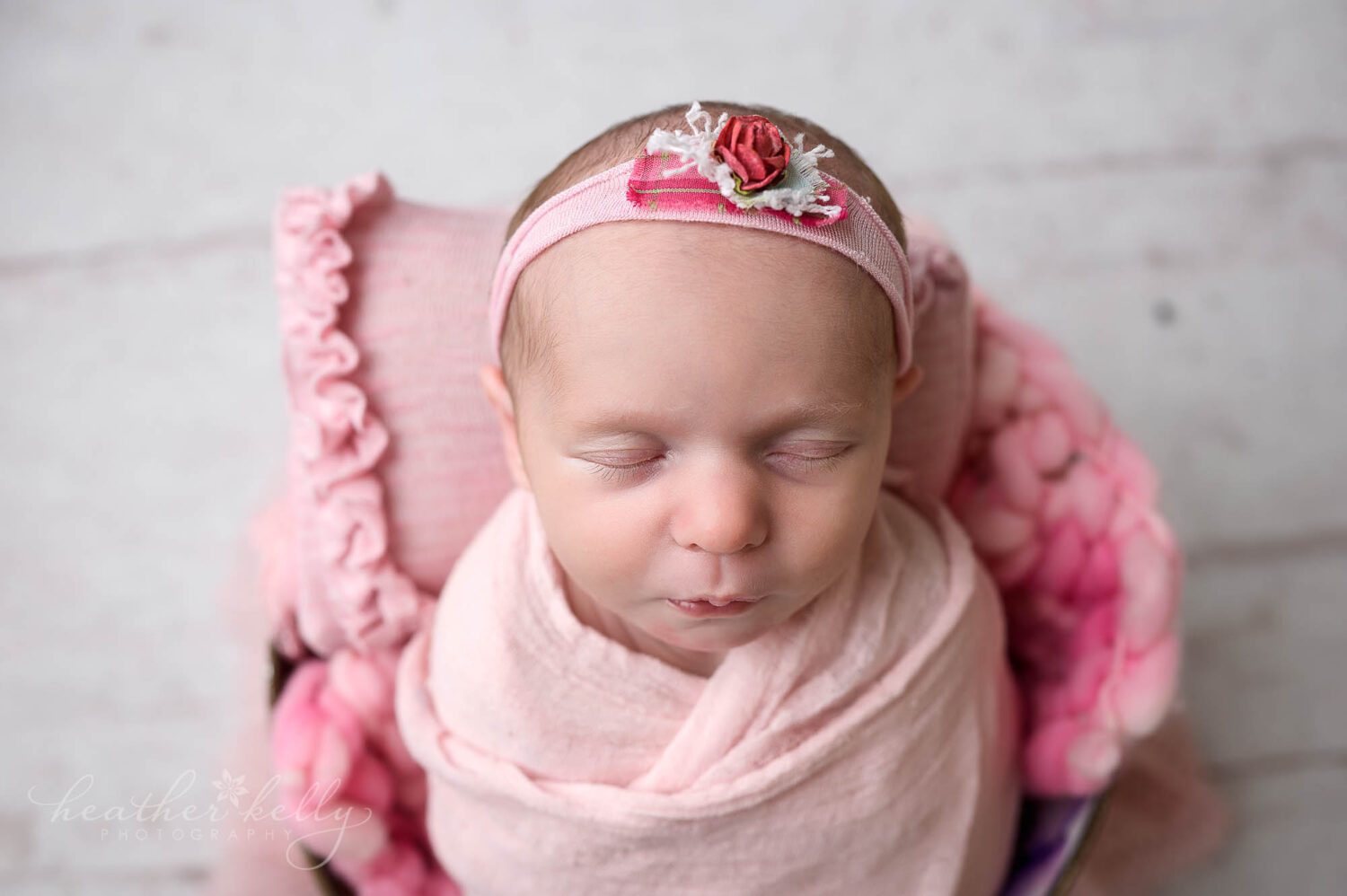Newborn photo of a newborn girl. She is wrapped in pink and is set inside a bucket. There is a small pink pillow behind her head. 