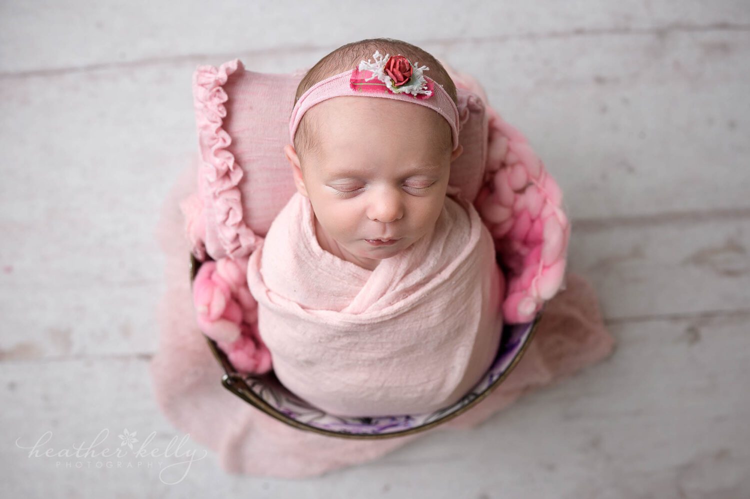A newborn girl during a photography session in CT. She is wrapped in pink. There is a pink small pillow behind her head. She is set inside a bucket. 