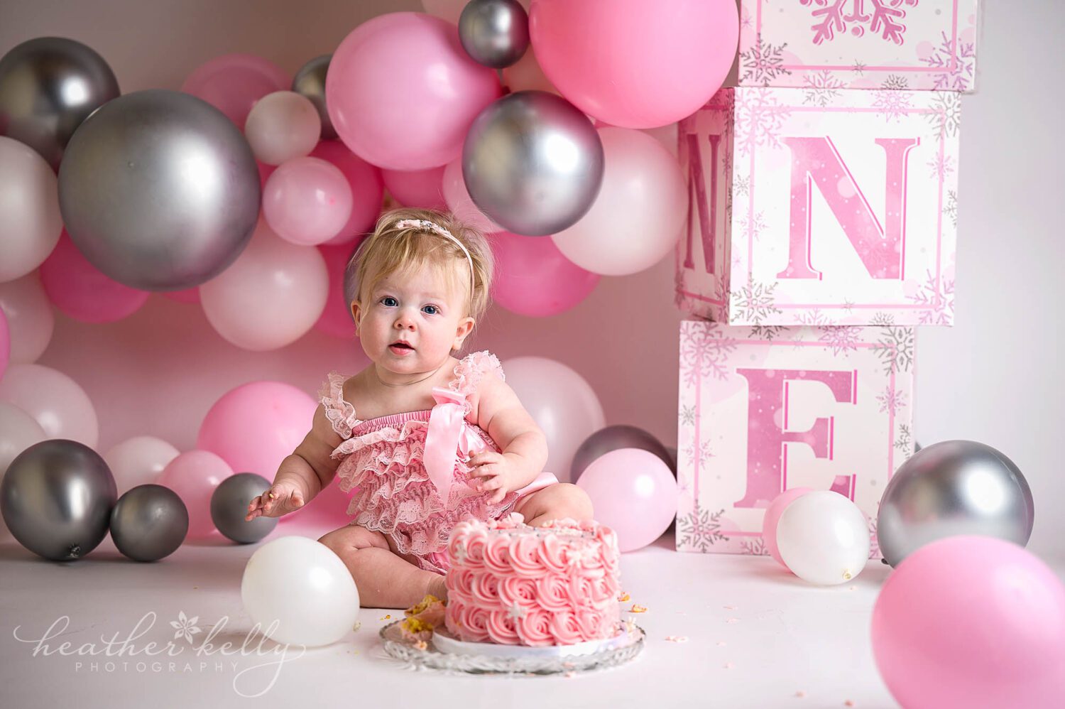 a portrait of a one year old girl with a pink cake, pink outfit, pink and gray balloons during her cake smash session in sandy hook ct. 
