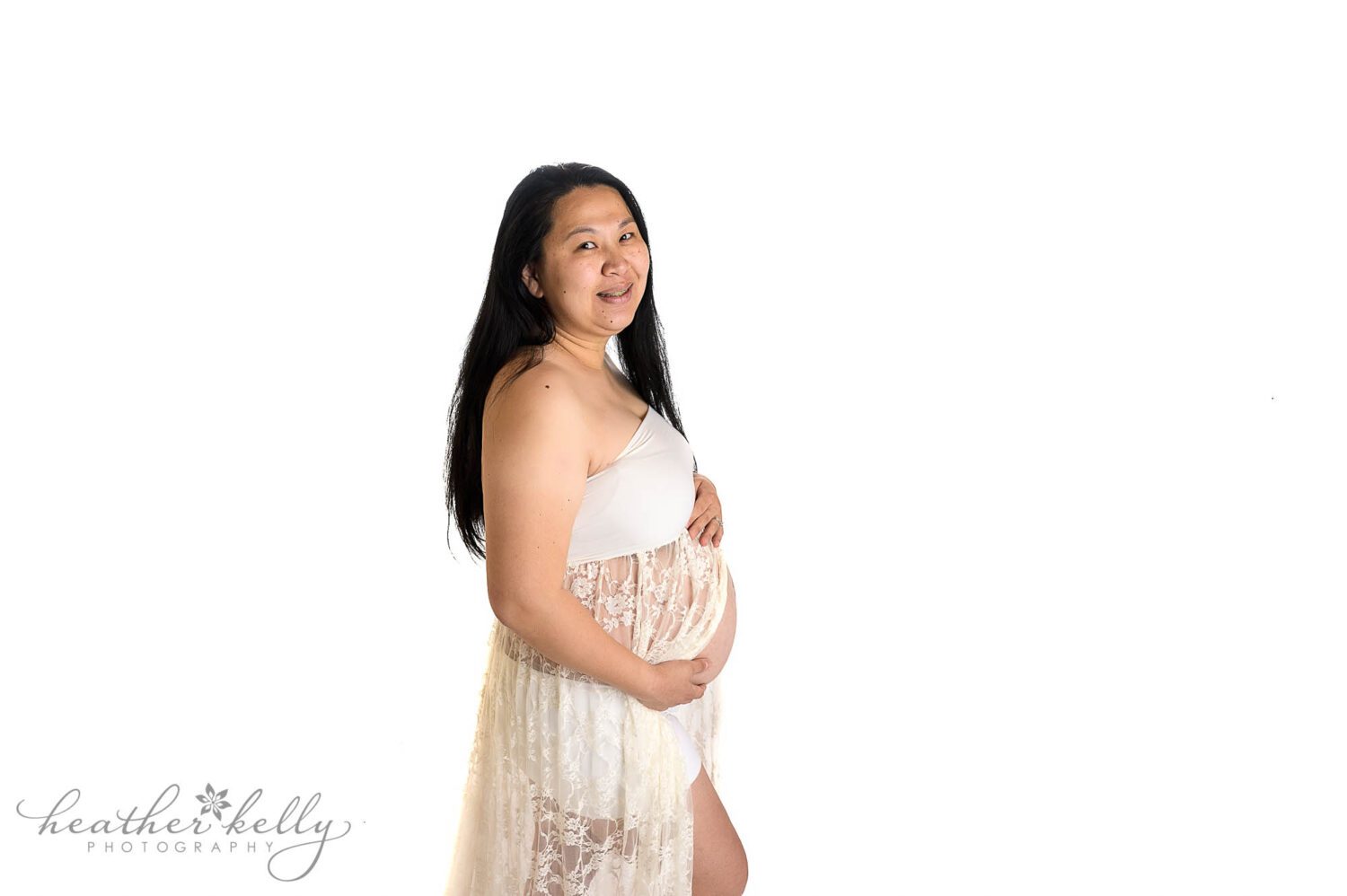 a mom to be wearing a white lace maternity gown during her maternity photography session. There is a high key white background. 