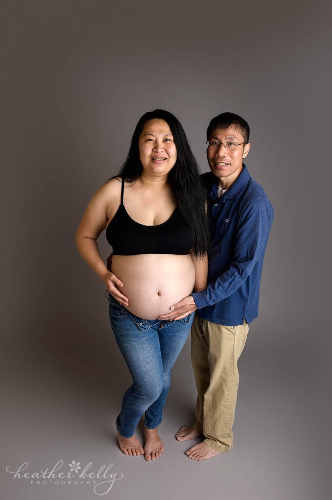 A maternity image with mom and dad. Dad is holding mom's belly. There is a gray background.  Danbury maternity photographer