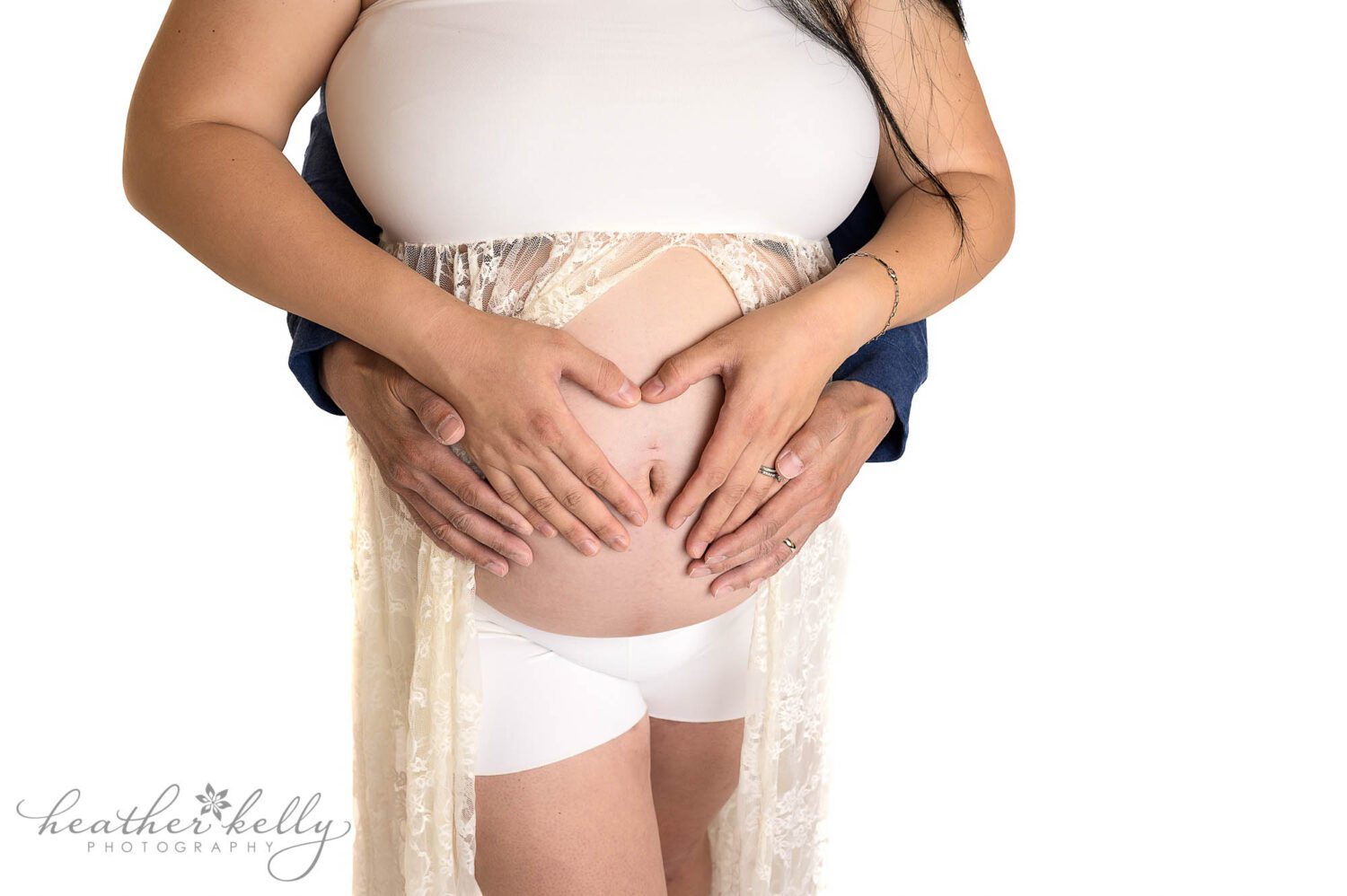 a mom to be wearing a white lace maternity gown during her maternity photography session. There is a high key white background. It is a close up image of mom's belly with her and dad's hands in a heart around her belly button. 