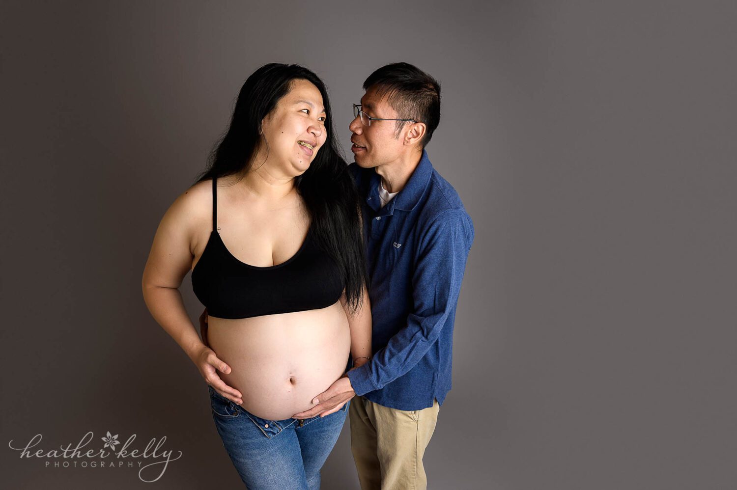 a man and woman are looking at each other. The woman is pregnant and wearing unbuttoned jeans and a black bra. The man's hand is holding the bottom of her belly. 