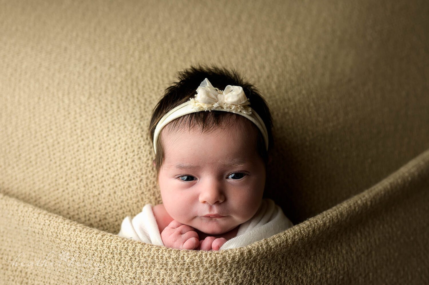 a newborn lays awake on a tan backdrop staring at the camera during her newborn photography session. 